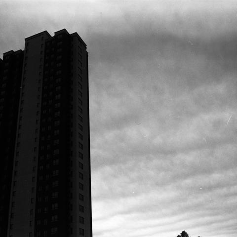 Lucky SHD 400 (6x6)

English Alt Text:
A dramatic black-and-white image shows a tall building silhouetted against a cloudy sky. The structure has many windows and looms on the left side of the frame. The sky is filled with layered clouds, creating a moody, textured backdrop. Tree tops appear in the lower right corner, adding contrast between nature and architecture. The photo emphasizes vertical scale and atmospheric tension.
中文替代文字：
这是一张黑白照片，画面左侧是一栋高楼的剪影，背景是层层叠叠的云层。建筑物窗户众多，显得高耸入云。右下角可见树梢，为画面增添…