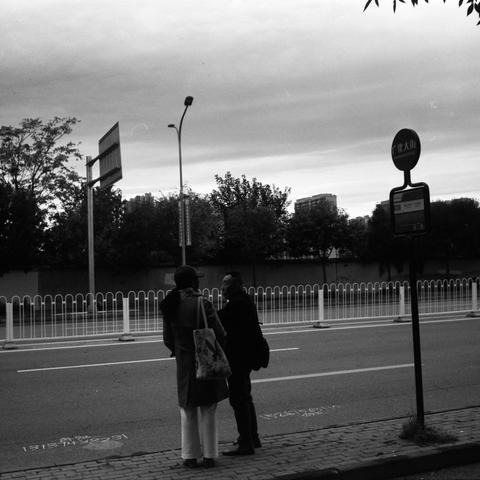 Lucky SHD 400 (6x6)

English Alt Text:
A black-and-white photo captures two individuals standing near a bus stop sign labeled “Guanghua Street” in Chinese. One person faces the sign, while the other looks down the road. The sidewalk is bordered by a white fence separating it from the street. Trees and distant buildings line the background. The pavement bears a safety warning in Chinese: “Please pay attention to safety.” The overcast sky and monochrome tones evoke a quiet, contemplative urban mo…