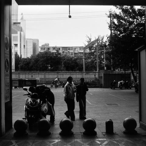 Lucky SHD 400 (6x6)

English Alt Text:
A black-and-white photo taken from inside a building looks out onto an urban courtyard. Spherical bollards line the entrance, and a fire extinguisher sits on the ground to the right. Three people are visible: one interacts with a scooter, while two others walk together. The background includes a fence, trees, and a mix of mid-rise and high-rise buildings. The scene captures everyday city life with quiet intimacy.
中文替代文字：
这是一张黑白照片，从室内望向城市庭院。入口处排列着几个球形路障，右侧地…