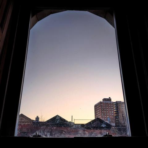 Looking through an arched window forty minutes after sunrise the cloudless sky fades from palest yellow at the horizon to steel blue up above. Pointed roofs of Harlem brownstones are silhouetted across the street, and a taller apartment building can be seen in the distance. The window is grimy on the bottom.