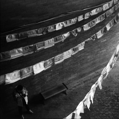 Lucky SHD 400 (6x6)

English Alt Text:
A black-and-white photo taken from above shows a narrow alley with a brick floor and wall. A person stands near a bench, looking down at their hands. Above them, several rows of rectangular cloth flags—resembling Tibetan prayer flags—are strung diagonally across the space. The flags add a sense of movement and cultural texture. The scene feels quiet and introspective, with the solitary figure framed by the geometric lines of the alley and the soft flutter …