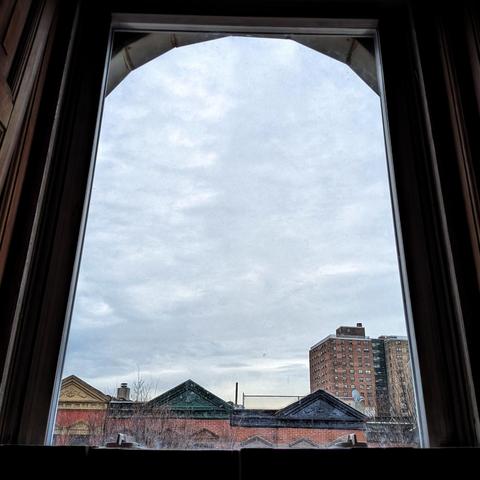 Looking through an arched window ninety minutes after sunrise the sky is filled with dense light gray clouds. Pointed roofs of Harlem brownstones with red brickwork are across the street, and a taller apartment building can be seen in the distance.