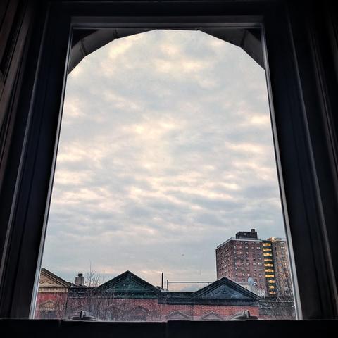 Looking through an arched window eleven minutes before sunrise the sky is filled with dense, lumpy gray and white clouds. Pointed roofs of Harlem brownstones with red brickwork are across the street, and a taller apartment building can be seen in the distance.