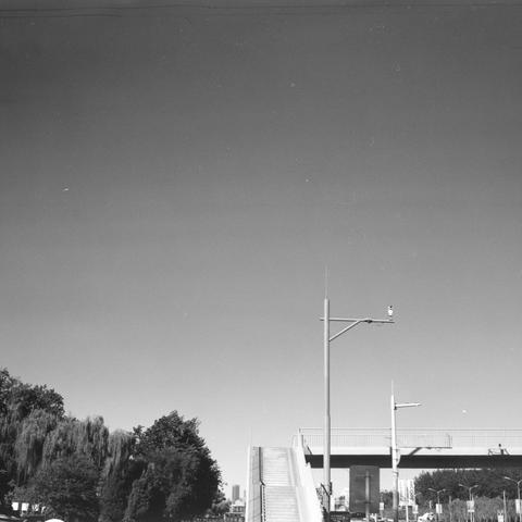 Lucky SHD 400 (6x6)
English:
This black-and-white photograph captures an urban scene under a clear sky. In the foreground, there are trees with dense foliage, and a pedestrian staircase leading up to an elevated walkway or bridge. The staircase is flanked by a tall streetlight. The bridge extends horizontally across the image, supported by pillars. The overall atmosphere is calm, with a focus on the architectural elements and the contrast between the sky and the structures.
Chinese (中文):
这张黑白照片…