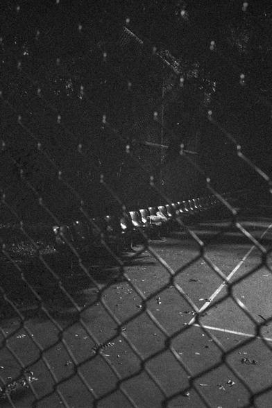 Ilford HP5 Plus 400 (FF)

English:
This black-and-white photograph captures a daytime scene at a sports field or park. A row of empty bleacher seats is visible in the foreground, aligned along a chain-link fence. The fence is illuminated by day light, casting dramatic shadows and highlights. The background is dark, with silhouettes of trees and foliage, creating a stark contrast between the lit area and the shadows.
Chinese (中文):
这张黑白照片拍摄于日间的运动场或公园。前景是一排空荡的看台座椅，沿着铁丝网围栏排列。围栏被明亮的日光照亮，投射出戏剧性的阴影和高光…