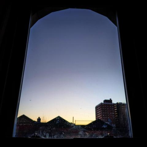 Looking through an arched window at the moment of sunrise the sky fades from pale orange at the horizon to deep blue up above. Pointed roofs of Harlem brownstones are silhouetted across the street, and a taller apartment building can be seen in the distance.