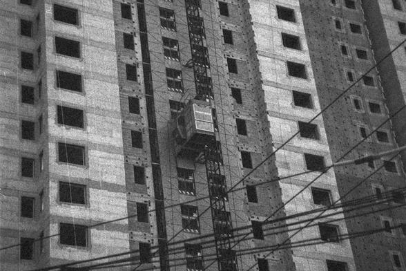 Ilford HP5 Plus 400 (FF)

English Alt Text: A black-and-white photo captures a high-rise building under construction. The concrete structure has multiple rectangular window openings, many without glass. A construction elevator is attached to a vertical scaffolding in the center, used for transporting materials and workers. Electrical cables run horizontally across the foreground. The grainy texture adds a documentary or vintage feel.

中文替代文字： 这是一张黑白照片，展示一栋正在施工的高层建筑。混凝土结构上有多个矩形窗洞，许多尚未安装玻璃。建筑中央有一…