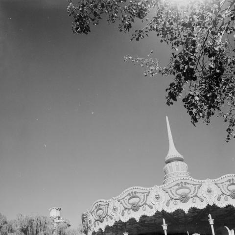 Lucky SHD 400 (6x6)

English Alt Text:
A black-and-white photograph showing the upper portion of a carousel with ornate decorations and a pointed spire. Tree branches partially obscure the top right corner. The sun shines brightly, creating a lens flare and casting light across the scene. The sky is clear, and more trees and a structure are visible in the background. The image evokes nostalgia and whimsy, reminiscent of amusement parks and childhood memories, with a dreamy and serene atmosphere…