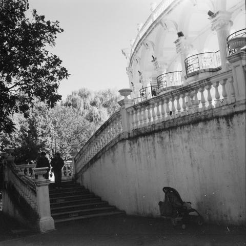 Lucky SHD 400 (6x6)

English Alt Text:
A black-and-white photo of a grand outdoor staircase leading to a classical-style pavilion. The staircase has ornate balustrades and wide stone steps. Two people walk up the stairs, silhouetted against the light. A stroller sits unattended near the bottom right corner. The pavilion features arches and columns, suggesting historical or public architecture. Dense trees form a leafy backdrop, and the lighting creates strong contrasts between shadow and sunlig…