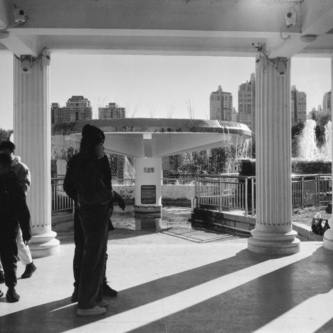 Lucky SHD 400 (6x6)

English Alt Text:
A black-and-white photo of a circular fountain with water jets shooting upward, located under a pavilion supported by classical columns. A railing surrounds the fountain. Several people walk or stand nearby, some in shadow. Tall buildings rise in the background, indicating an urban setting. Sunlight casts long shadows across the scene. A sign with Chinese characters is visible near the base of the fountain, suggesting the location is in a Chinese-speaking …