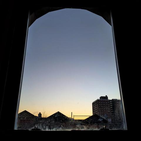 Looking through an arched window at the moment of sunrise the sky fades from pale orange at the horizon to deep blue up above. Pointed roofs of Harlem brownstones are silhouetted across the street, and a taller apartment building can be seen in the distance.