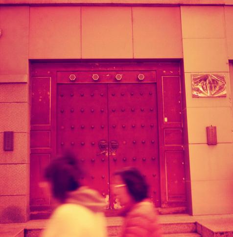 Harman Red 125 (6x6)

English Alt Text:
A large traditional wooden door with metal studs and ornate lion head knockers is set into a stone facade. The door has symmetrical rectangular panels and five hexagonal decorations above it. Two plaques flank the door, one with Chinese characters. Two blurred figures walk past in the foreground, adding motion to the otherwise static architectural scene. The image has a reddish-pink tint, giving it a stylized, vintage feel.
中文替代文本：
一扇大型传统木门嵌有金属铆钉和精致的狮头门环，…
