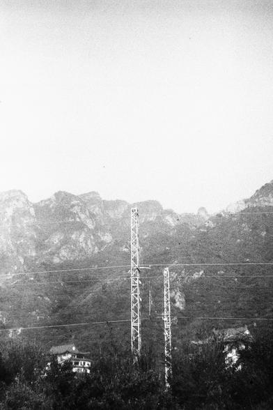 ERA 100 (FF)

English Alt Text:
A black and white image of a mountainous landscape with rugged terrain in the background. Two tall metal transmission towers dominate the foreground, with power lines stretching horizontally across the sky. At the base of the towers, rooftops of buildings peek through the vegetation. The scene contrasts natural majesty with industrial presence, highlighting how infrastructure intersects with untouched land. The monochrome effect adds drama and starkness to the co…