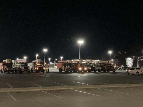 A mostly empty parking lot at night, with 8 bucket trucks and their crews milling about.