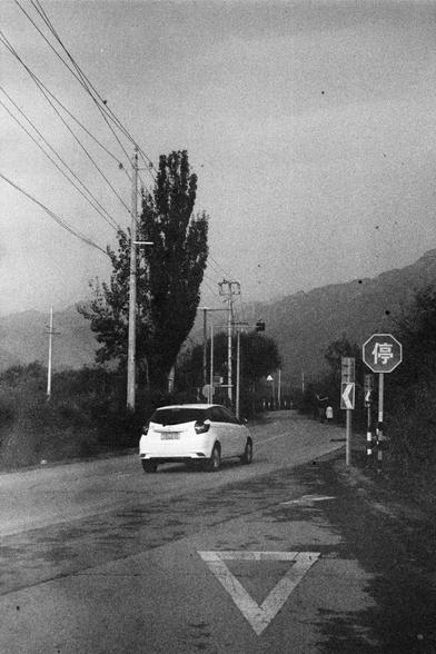 ERA 100 (FF)

English Alt Text:
A black-and-white photo of a rural road curving through a forested area. A white car drives along the road, which is lined with tall trees and utility poles. Power lines stretch overhead. Road signs include a stop sign with the Chinese character “停”, a yield symbol painted on the road, and a directional arrow indicating a curve. Mountains are visible in the distance. The image has a grainy, nostalgic feel.

中文替代文字：
这是一张黑白乡村道路照片，一辆白色汽车行驶在弯曲的道路上，道路两旁是高大的树木和电线杆，电线横跨…
