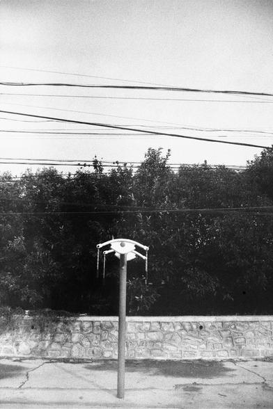 ERA 100 (FF)

English Alt Text:
A surreal black-and-white image showing a sculptural pole in a street setting. At the top of the pole is an eye-shaped structure with two horizontal rods extending outward, each with chains hanging down. Behind the pole is a stone wall and dense foliage. Above the trees, multiple power lines stretch across the sky. The ground is paved and shows signs of wear. The eye sculpture evokes themes of surveillance or awareness.

中文替代文字：
这是一张黑白超现实风格的街景照片，画面中央是一根雕塑杆。杆顶是一个眼…