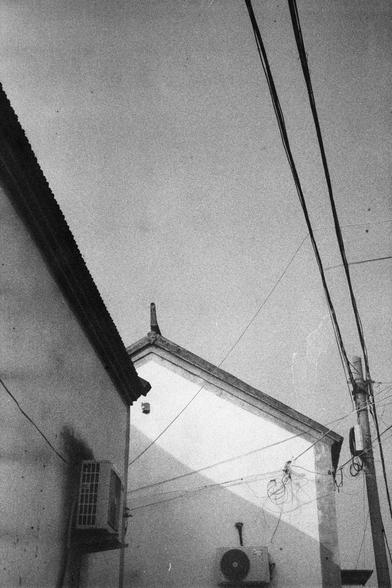 ERA 100 (FF)

English Alt Text: A grayscale image features the upper portions of two buildings with traditional-style rooftops. Both buildings have air conditioning units mounted on their exterior walls. A cluster of electrical wires connects to the building on the right, and several power lines run diagonally across the sky. A utility pole is visible on the right edge. The architectural style and infrastructure suggest an older urban setting, possibly in East Asia, with a mix of tradition and …