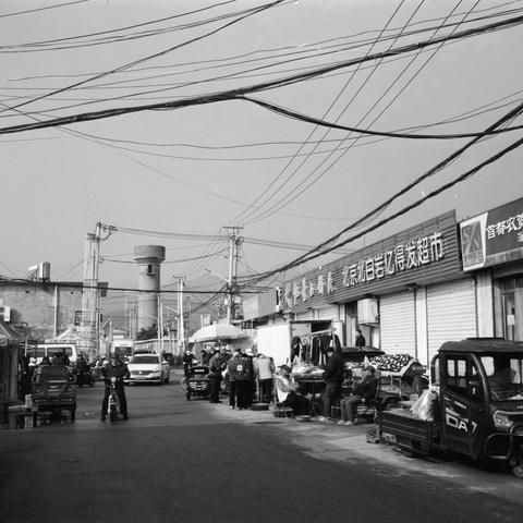 Lucky SHD 400 (6x6)

English Alt Text:
A black and white street scene in a bustling commercial area of a Chinese city. Storefronts with Chinese signage line the right side, including “北京悦百信亿得发超市” and “首都农资.” People gather around market stalls, and vehicles including a three-wheeled truck labeled “DA7” move through the street. Overhead wires crisscross above, and a cylindrical water tower rises in the background. The image captures everyday urban life with rich detail and movement.
中文替代文本：
一张黑白街…