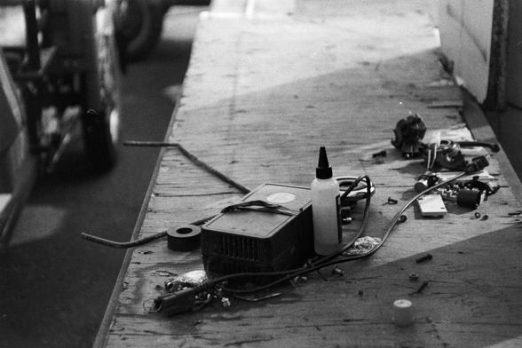 NEOPAN SS 100 (FF)

English Alt Text:
A black-and-white photo of a cluttered workbench filled with tools and electronic components. Items include a squeeze bottle, a vented electronic device with wires, a roll of tape, screws, bolts, and disassembled mechanical parts. The background shows a vehicle and a wall, suggesting a garage or workshop setting. The scene conveys hands-on technical work in progress or paused.
中文替代文字：
这是一张黑白照片，展示了一个杂乱的工作台，上面堆满了工具和电子零件。可见物品包括尖嘴塑料瓶、有通风孔和电线的电子设备、胶带卷、螺丝、螺栓以及拆解的…