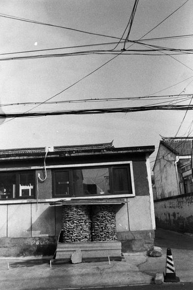 NEOPAN SS 100 (FF)

English Alt Text:
A black-and-white photo showing two large cylindrical stacks filled with corn cobs in front of a flat-roofed building. The building has several windows, an air conditioning unit, and hanging cables. A striped traffic cone sits on the ground amid debris. Overhead, power lines crisscross the sky. The image captures everyday urban life with elements of food storage and infrastructure.
中文替代文字：
这是一张黑白照片，画面中有两大圆柱形玉米堆，位于一座平顶建筑前。建筑有多个窗户、一个空调机和垂挂的电缆。地面上有一个黑白条纹的交通锥和一…