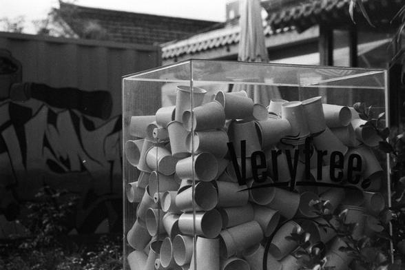 NEOPAN SS 100 (FF)

English Alt Text:
A black-and-white photo showing a transparent rectangular container filled with numerous disposable cups. The word “Veryfree.” is printed boldly on the container’s side. Behind it, a graffiti mural decorates the wall, and a building with traditional roof tiles is visible. An umbrella suggests an outdoor setting. The image may comment on consumerism or environmental waste, with the discarded cups confined in a clear box. The monochrome palette adds a reflect…