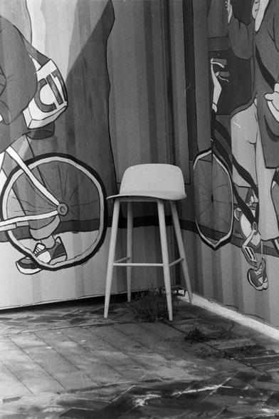 NEOPAN SS 100 (FF)

English Alt Text:
A monochrome photo of a corner where two tiled walls meet. The walls are painted with a mural of cartoon-style people riding bicycles. The figures are playful and stylized, adding whimsy to the scene. In the corner sits a modern bar stool with a curved seat and four legs. The tiled floor has small patches of grass growing between the cracks and at the base of the walls. The image blends real objects with artistic elements, creating a surreal, urban atmosphe…
