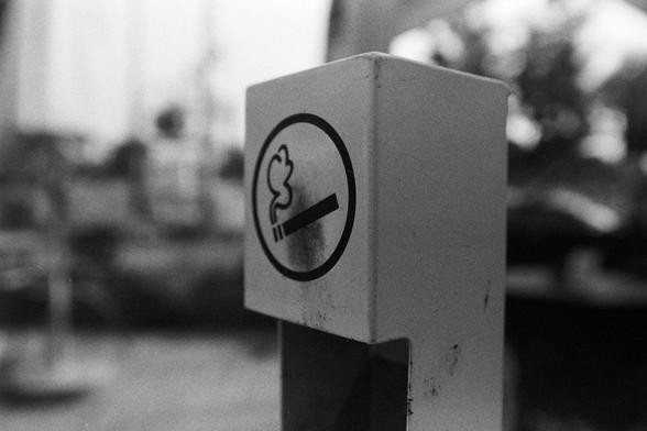 NEOPAN SS 100 (FF)
English Alt Text:
A close-up black and white photo of a public smoking area sign. The sign features a cigarette with smoke inside a circle, indicating smoking is allowed. The surface is worn, with dark smudges and burn marks. The background is blurred, showing trees and structures. The image highlights the wear and usage of designated smoking zones.

中文替代文字：
这是一张黑白特写照片，展示了一个公共吸烟区标志。标志上画有一支冒烟的香烟，圈在圆形中，表示此处允许吸烟。标志表面有明显的污渍和烧痕。背景模糊，可见树木和建筑结构。画面突出了吸烟区长期使用后的磨损痕迹。