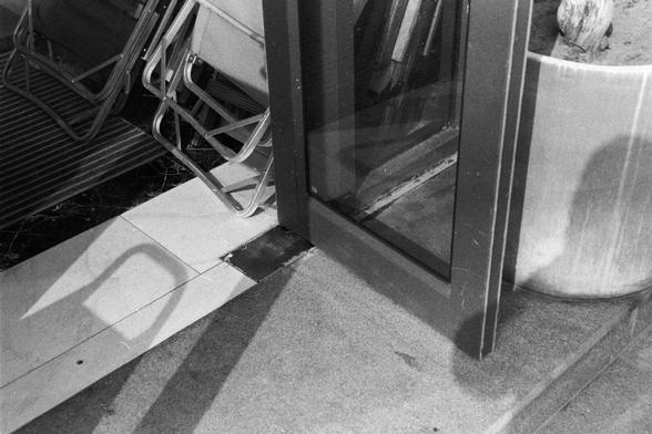 NEOPAN SS 100 (FF)
English Alt Text:
A black and white photo of a partially open glass door leading into a building. Metal folding chairs are stacked against the wall to the left. The floor has a mix of textures: a mat, black marble tiles, and light-colored tiles. A large planter with driftwood sits on the right. Shadows from the door and chairs stretch across the floor, adding depth and contrast.

中文替代文字：
这是一张黑白照片，展示了一扇半开玻璃门通向建筑内部。左侧墙边堆放着几把金属折叠椅。地面由多种材质组成，包括地垫、黑色大理石瓷砖和浅色瓷砖。右侧有一个大型花盆，里面放着一块漂流木。…