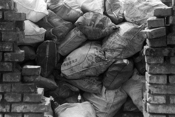 NEOPAN SS 100 (FF)

English Alt Text: A gritty black-and-white photo showing a pile of large sacks stacked behind a partially built or broken brick wall. The sacks are made of woven plastic or fabric and bear Chinese text, possibly indicating industrial or construction materials. Some sacks are dirty and worn. A few glass bottles lie at the bottom of the pile. The scene suggests a utilitarian or industrial setting, with textures of rough brick, fabric, and debris contributing to a raw, document…