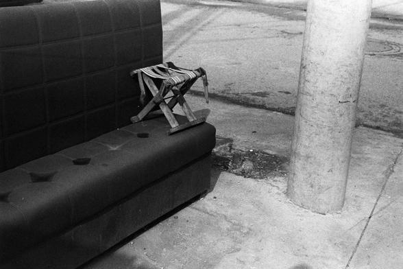 NEOPAN SS 100 (FF)

English Alt Text: A black-and-white street scene featuring a stained, tufted sofa placed on a paved surface. On top of the sofa sits a small, foldable wooden stool with frayed fabric straps, appearing aged and possibly broken. To the right stands a large cylindrical concrete utility pole. The ground shows cracks and patches, with a small exposed dirt area near the pole’s base. The background includes faint street markings and a manhole cover. The image conveys urban decay an…