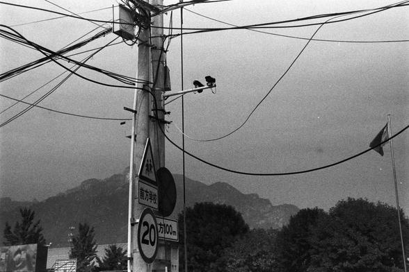 NEOPAN SS 100 (FF)

English Alt Text: A black-and-white photo of a utility pole densely packed with surveillance cameras, overhead wires, and traffic signs. A triangular sign shows two children walking, indicating a school zone. Below it, a rectangular sign in Chinese reads “School Ahead.” A circular sign displays “20,” marking a speed limit of 20 km/h. Another sign reads “100 meters ahead.” In the background are trees, mountains, and a flagpole with the Chinese national flag. The image reflect…