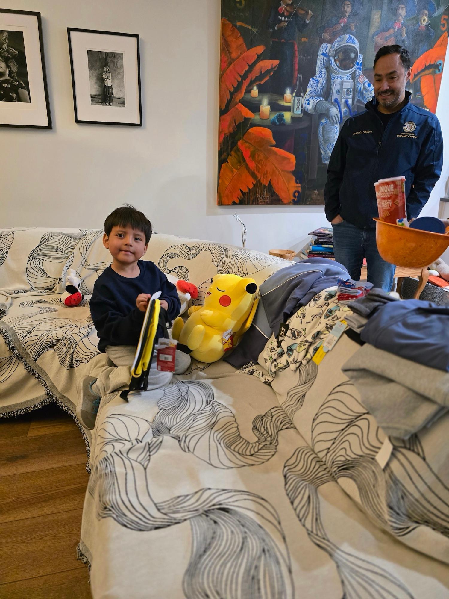 Liam sitting on his couch with some of his belongings including a plush Pikachu. He has a little smile. Congressman Castro stands behind the couch looking at him with a smile.
