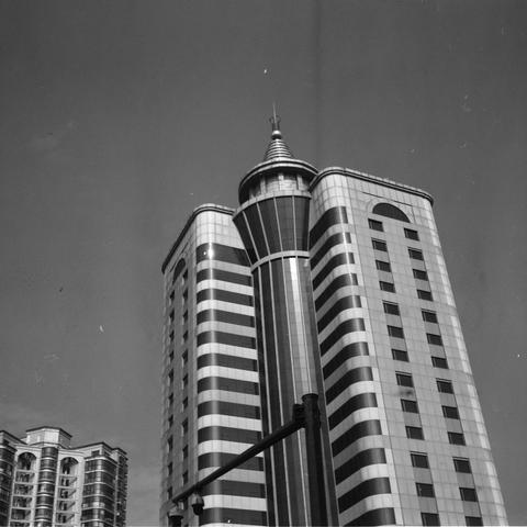 Lucky SHD 400 (6x6)

English Alt Text:
A black-and-white photograph of a futuristic high-rise building. The central tower features a cylindrical shape that narrows into a pointed spire, resembling a rocket or needle. The facade is patterned with alternating horizontal stripes in light and dark tones, giving it a sleek, modern appearance. Windows are arranged in a uniform grid across the surface. To the left, another building with balconies is partially visible. A traffic light pole stands in th…