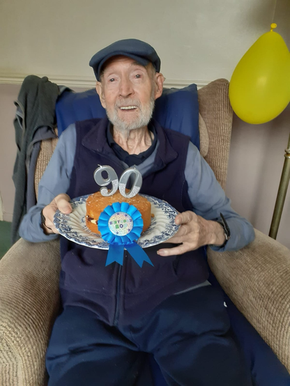 Lee's dad, sitting in a cozy chair, wearing a hat and holding a cake adorned with a blue ribbon and candles indicating it's his 90th birthday. You can absolutely see the resemblance between Lee and his dad.