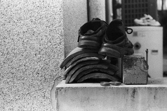 NEOPAN SS 100 (FF)

English Alt Text:
A monochrome photograph showing a pair of athletic shoes placed atop a stack of curved roof tiles arranged in a semi-circle on a concrete surface. Beside them is a weathered concrete block with a metal rod protruding. In the blurry background, another pair of shoes rests on a white cabinet. The image captures urban textures and everyday objects with a nostalgic tone.
中文替代文字：
一张黑白照片，展示一双运动鞋放在一堆弯曲的屋顶瓦片上，瓦片呈半圆形排列在水泥地面上。旁边是一个老旧的水泥块，插着一根金属杆。模糊的背景中，另一双鞋放在一个白色柜子上。…