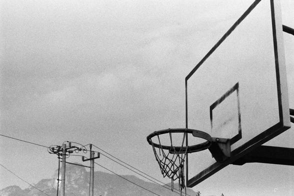 NEOPAN SS 100 (FF)

English Alt Text:
A black-and-white photo of an outdoor basketball hoop mounted on a rectangular backboard with a marked square target. The hoop has a net and stands against a cloudy sky. In the background, utility poles and power lines stretch across the scene, with faint hills or mountains visible in the distance. The image evokes a quiet, nostalgic mood, emphasizing simplicity and open-air recreation.
中文替代文字：
一张黑白照片，展示一个户外篮球框，安装在一个带有方形标记的矩形篮板上。篮筐挂着网，背景是多云的天空。远处有电线杆和电线横穿画面…