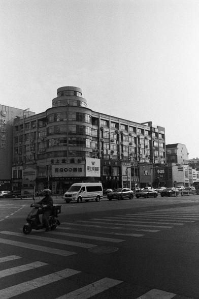 NEOPAN SS 100 (FF)

Image 3 – English Alt Text
A black‑and‑white street scene featuring a rounded‑corner multi‑story building with many windows. The ground floor hosts several storefronts with Chinese signage, including shops and restaurants. Cars, a white van, and a person on a scooter move through the street. A pedestrian crosswalk spans the foreground. The image captures a busy urban environment.
Image 3 – 中文替代文字
黑白城市街景。一栋圆角多层建筑占据画面，外墙布满窗户。底层是带中文招牌的商铺，如眼镜店和餐馆。街上有汽车、白色货车和骑电动车的人。前景是人行横道，呈现典型的城…