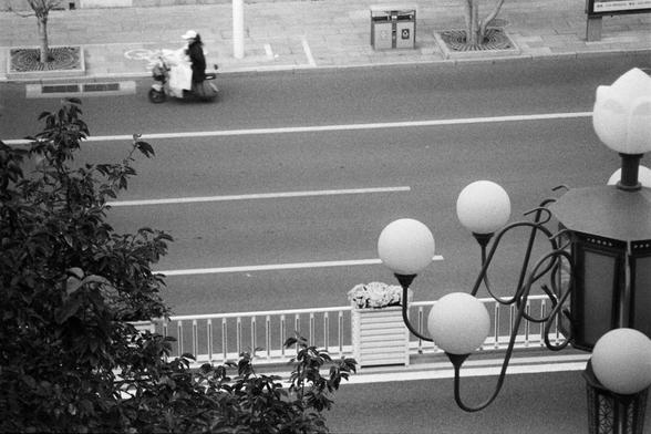 NEOPAN SS 100 (FF)

Image 1 – English Alt Text
A black‑and‑white overhead view of a wide, mostly empty street. A single person rides a scooter in the center lane, carrying several large bags or bundles stacked behind them. The road markings are clean and geometric. On the sidewalk to the left, there are two trash bins, a bicycle‑lane symbol, a tall pole, and several young trees planted in square grates. In the foreground, an ornate streetlamp with multiple spherical bulbs stands beside a plante…