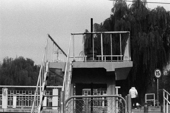 NEOPAN SS 100 (FF)

Image 1 — English Alt Text
A black‑and‑white photo of a small raised platform made of metal, with railings and two staircases leading up from opposite sides. A tall vertical pole stands at the center of the platform. Behind it are large trees with long, drooping branches. To the right, a circular road sign displays a weight limit of “15t.” A person in a light‑colored coat stands near the sign. The scene appears outdoors, with a contrast between the industrial structure and t…