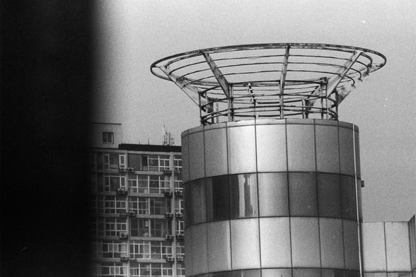 NEOPAN SS 100 (FF)

Image 2 — English Alt Text
A black‑and‑white photograph showing a modern cylindrical building covered in reflective glass panels. At the top is a circular metal framework. Behind it stands a rectangular building with many windows and several air‑conditioning units attached to the exterior. The left side of the image is partially obscured by a dark vertical shadow or object. The composition emphasizes contrasting architectural shapes.
Image 2 — 中文替代文字
一张黑白照片，主体是一座圆柱形的现代建筑，外墙由…