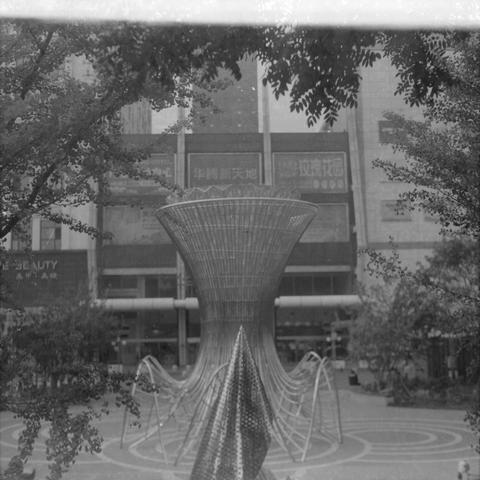Ilford HP5 Plus 400 (6x6)

English Alt Text
A black‑and‑white image of a large metal public sculpture in a city plaza. The central structure is a tall, funnel‑shaped form made of intersecting metal rods, surrounded by curved wave‑like metal elements. Buildings with Chinese signage and trees frame the scene. The ground features circular patterns.
Chinese Alt Text
一张黑白照片，展示城市广场中的大型金属公共艺术装置。中心是由金属杆构成的漏斗状结构，周围环绕着弧形的波浪状金属线条。背景有带中文招牌的建筑和树木，地面上有圆形图案。