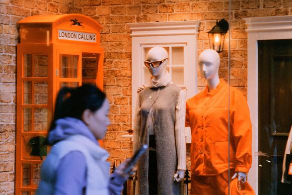 Harman Phoenix 200 II (FF)

English Alt Text:
Two mannequins stand in a storefront display. One wears a long gray sleeveless coat layered over a cream turtleneck sweater, accessorized with sunglasses. The other mannequin is dressed in a bright orange jacket and matching skirt, holding an orange handbag. Behind them is a brick wall with white-framed windows and a lantern-style light fixture. To the left, a vivid orange British-style telephone booth with the words “LONDON CALLING” and a spider gr…