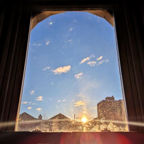 Looking through an arched window half an hour after sunrise the sun is just above the buildings, making a dramatic lens flare that bisects the frame horizontally at the bottom with a curved yellow line and multi-colored petals below. The sky is light blue with scattered white clouds. Pointed roofs of Harlem brownstones are silhouetted across the street, and a taller apartment building can be seen in the distance. The window is grimy at the bottom.