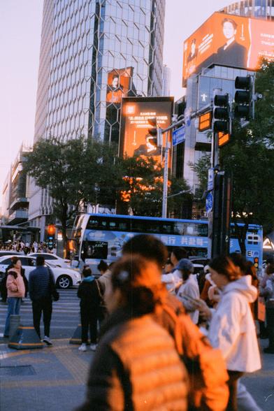 Harman Phoenix 200 II (FF)

English Alt Text:
A lively city street at dusk with crowds of pedestrians crossing. Tall glass buildings rise in the background, reflecting fading daylight. Bright digital billboards glow in red-orange tones, including one promoting cosmetics with the brand “KANS 韩束” and “TYAKASHA.” The illuminated signage contrasts with the dim sky, emphasizing digital media’s presence in urban life. Buses, cars, traffic lights, and the “Renmin Middle Rd” sign add order to the bustl…