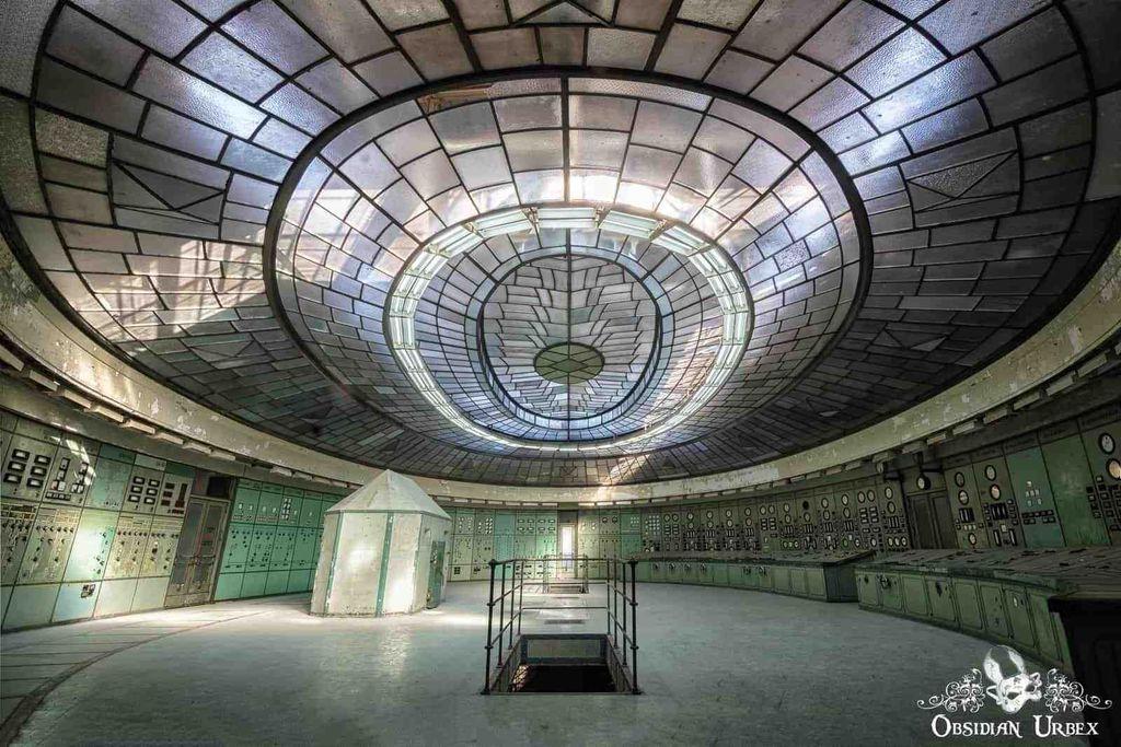 An ornate, abandoned power plant control room features a magnificent circular glass dome ceiling. Light fills the room, highlighting teal control panels lining the walls and a central white structure.