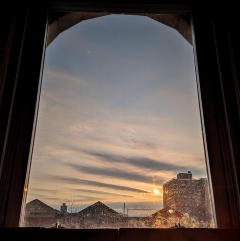Looking through an arched window fifty-three minutes after sunrise the orange sun is just above the buildings on the right and the slate blue sky is slashed with several dramatic lines of grey clouds above the horizon. Pointed roofs of Harlem brownstones are silhouetted across the street, and a taller apartment building can be seen in the distance. The window is grimy on the bottom.