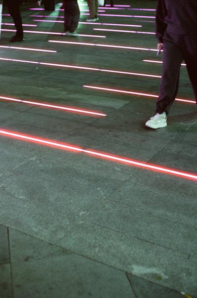 CineStill 800T (FF)

Image 3 – English Alt Text
A paved walkway with embedded horizontal red LED light strips glowing at night. Several people walk across the area, but only their legs and feet are visible. The red lights contrast with the gray stone surface, creating a modern, graphic pattern that guides movement or adds decorative interest.
Image 3 – 中文替代文字
一条铺石步道上嵌有水平红色 LED 灯带，在夜间发光。几个人从灯带上方走过，只能看到他们的腿和脚。红光与灰色石面形成鲜明对比，呈现出现代感强烈的视觉效果。