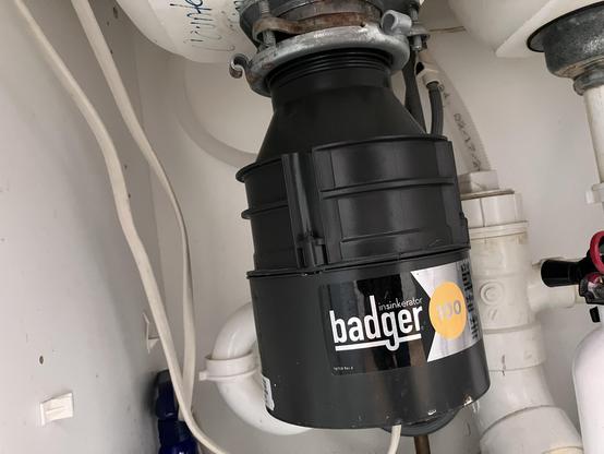 An under-sink garbage disposal unit labeled "Badger 100." It is black with a circular design and attached to plumbing beneath a sink.