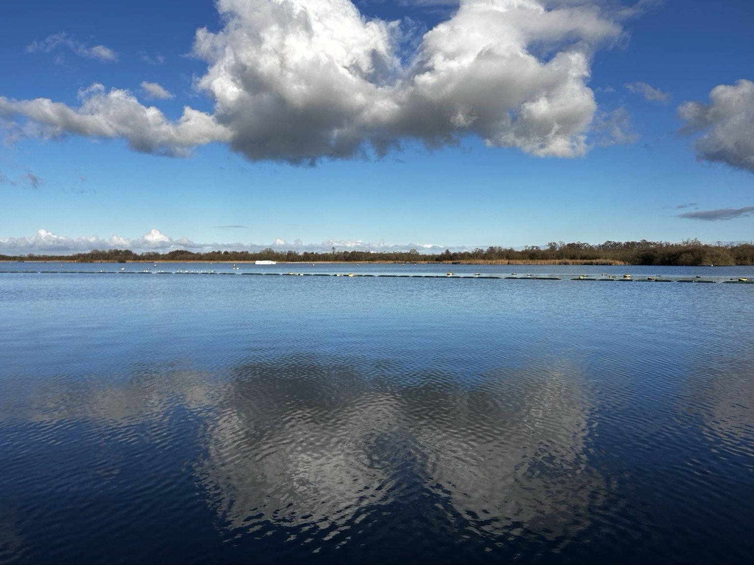 White fluffy clouds in a bright blue sky reflect on the clear still water of Barton Broad