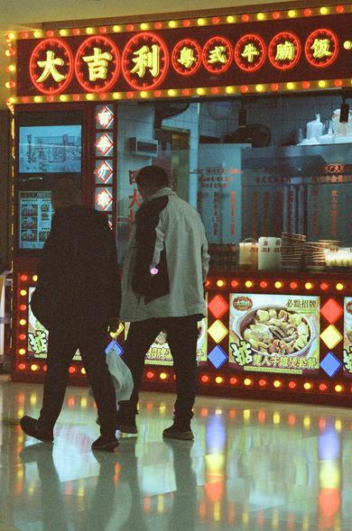 CineStill 800T (FF)

English Alt Text
A brightly lit food stall with red and yellow signage advertising Cantonese-style beef brisket rice. Illuminated menu boards show photos of dishes and promotional items. Two people walk past the stall, while the interior reveals kitchen equipment and stacked containers. The atmosphere is lively and colorful.
Chinese Alt Text
一个灯光明亮的餐饮摊位，红黄招牌宣传粤式牛腩饭。发光的菜单板展示多种菜品和促销信息。两个人从摊位前走过，内部可见厨房设备和堆放的容器。整体氛围热闹而多彩。